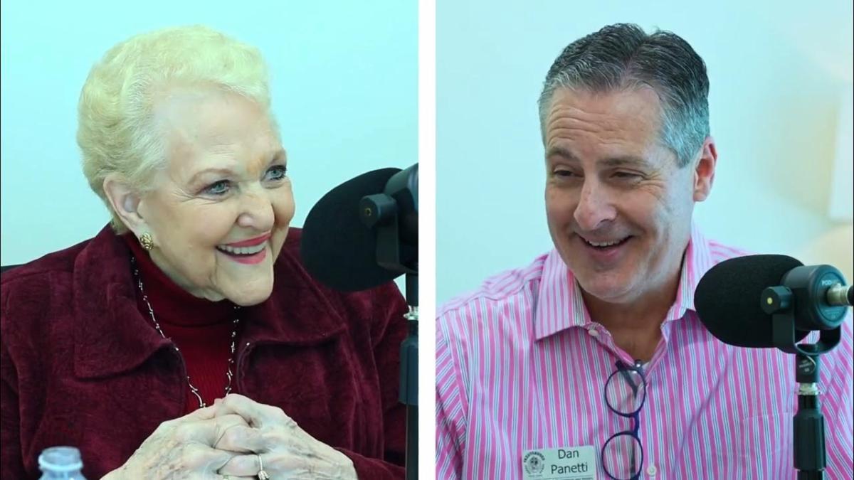 Two people sit across from each other at microphones in a recording setup, smiling warmly as they talk. One is an older woman wearing a red jacket, and the other is a man in a striped shirt with glasses hanging from his collar.